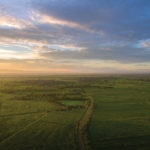 Aerial view of a vast, sunlit landscape with green fields, a winding road, and scattered trees. The sky above is filled with dramatic clouds, illuminated by the warm, golden hues of the setting sun on the horizon.