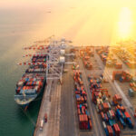 Aerial view of a busy shipping port at sunset, with large cranes loading cargo containers onto ships docked by the waterfront and stacks of colorful containers lined up along the docks.