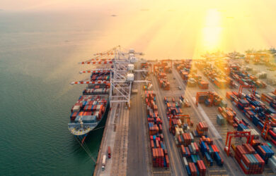Aerial view of a busy shipping port at sunset, with large cranes loading cargo containers onto ships docked by the waterfront and stacks of colorful containers lined up along the docks.