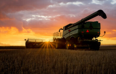 A combine harvester sits in a harvested field at sunset, with sun rays shining through its wheels and dramatic clouds in the sky.