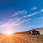A red tractor plows a vast, open field under a wide blue sky with scattered clouds. The sun is low on the horizon, casting a warm glow over the landscape and creating long shadows. Dust rises in the wake of the tractors path.