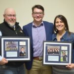 Three people smiling and holding framed photographs and certificates proudly represent Boonville FFA. The person on the left wears a black vest, the middle one dons a blue suit, and the person on the right sports a denim jacket, all standing against a plain wall.