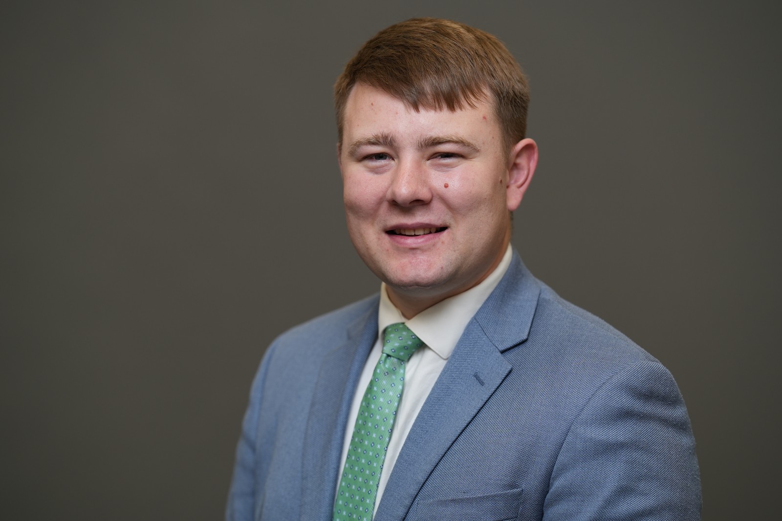 A young man in a light blue suit jacket, white shirt, and green polka dot tie smiles in front of a plain gray background.