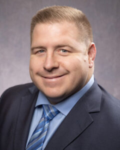 A person with short light brown hair, wearing a blue suit jacket, light blue shirt, and a striped blue tie. They are smiling and looking towards the camera against a plain background.