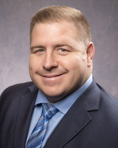 A person with short light brown hair, wearing a blue suit jacket, light blue shirt, and a striped blue tie. They are smiling and looking towards the camera against a plain background.