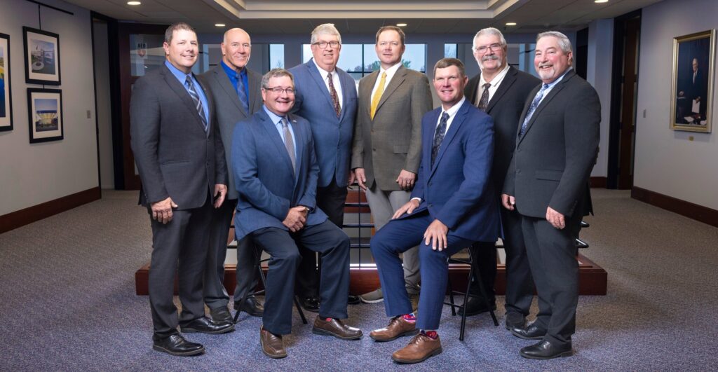 Eight men in suits pose for a formal group photo in a well-lit hallway with framed pictures on the walls and carpeted floors. Two men are seated on stools, while the others stand behind them, all smiling at the camera.