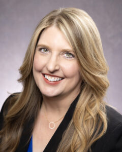 Portrait of Jennifer Bach Vice President of Break Time Operations with long blonde hair is smiling. She is wearing a black blazer over a blue top and a gold necklace with a circular pendant. The background is a soft gradient of gray and purple.