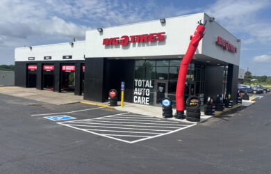 A Big O Tires auto care shop with service bays, a red inflatable tube man near the entrance, tire displays outside, and a parking lot with a wheelchair-accessible parking space.