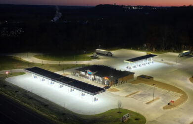 Aerial view of a Breaktime gas station with multiple fuel pumps and a convenience store, surrounded by an empty parking lot at dusk. A semi-truck is parked nearby. The sky is darkening, and the area is quiet.