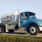 A blue and white propane delivery truck labeled MFA Oil Propane is parked on a rural road. The truck has large tires and a cylindrical tank. The background shows a grassy field under a clear blue sky.