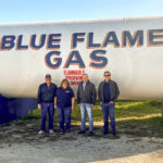Four people stand in front of a large propane tank labeled Blue Flame Gas with warnings about flammable propane and no smoking. The group poses outdoors on a sunny day, with grassy ground beneath them.
