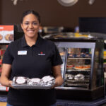 A smiling employee in a black uniform holds a tray with wrapped sandwiches in a convenience store, standing in front of a display case with pastries and a pizza and snacks sign.