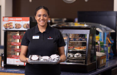 A smiling employee in a black uniform holds a tray with wrapped sandwiches in a convenience store, standing in front of a display case with pastries and a pizza and snacks sign.