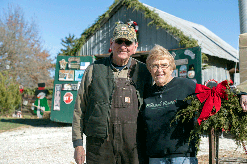 An elderly couple stands outside a rustic, decorated barn. The man wears a cap and overalls, while the woman is in a black sweatshirt. They smile at the camera, surrounded by festive greenery and bows. The sky is clear and blue.