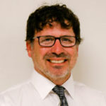 Man with glasses, dark curly hair, and a beard, smiling. Hes wearing a white shirt and a patterned tie, standing against a plain background.