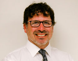 Man with glasses, dark curly hair, and a beard, smiling. Hes wearing a white shirt and a patterned tie, standing against a plain background.
