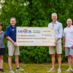 Four people stand outdoors holding a large check for $145,000 made out to Operation Homefront. The check is from the MFA Oil Charity Golf Scramble. Green trees are in the background.