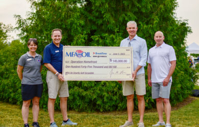 Four people stand outdoors holding a large check for $145,000 made out to Operation Homefront. The check is from the MFA Oil Charity Golf Scramble. Green trees are in the background.