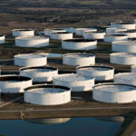Aerial view of numerous large, white, circular oil storage tanks arranged in rows on a sprawling industrial site. A small water body is visible in the foreground, with trees and landscape in the background.