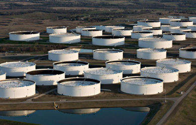 Aerial view of numerous large, white, circular oil storage tanks arranged in rows on a sprawling industrial site. A small water body is visible in the foreground, with trees and landscape in the background.