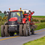 A red tractor with double rear wheels pulling farming equipment on a rural road. Rolling green fields and a barn are visible in the background. The sky is clear and blue.
