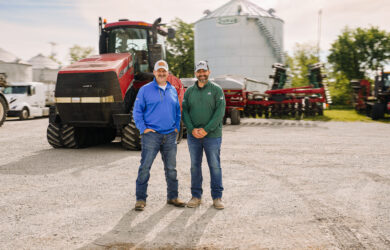 Drew and Colby Dixon stand smiling in front of a large red tractor and agricultural equipment on a farm, with a white grain bin and trees visible in the background under a bright, partly cloudy sky.