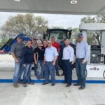 A group of nine people are standing together under a canopy at a fueling station with two pumps on either side. A blue tractor is visible in the background. They are smiling and wearing casual clothes, some with hats. Trees and vehicles are in the background.