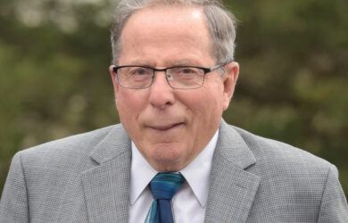 David Baker with glasses, wearing a gray suit, white shirt, and patterned blue tie, stands outdoors in front of blurred green foliage.
