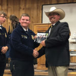A man in a cowboy hat shakes hands with a younger man in a blue jacket at an indoor event. The younger man holds a plaque, and they both smile. Other people are visible in the background.