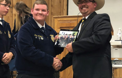 A man in a cowboy hat shakes hands with a younger man in a blue jacket at an indoor event. The younger man holds a plaque, and they both smile. Other people are visible in the background.