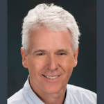 Smiling man with short white hair, wearing a light-colored collared shirt, set against a dark background.