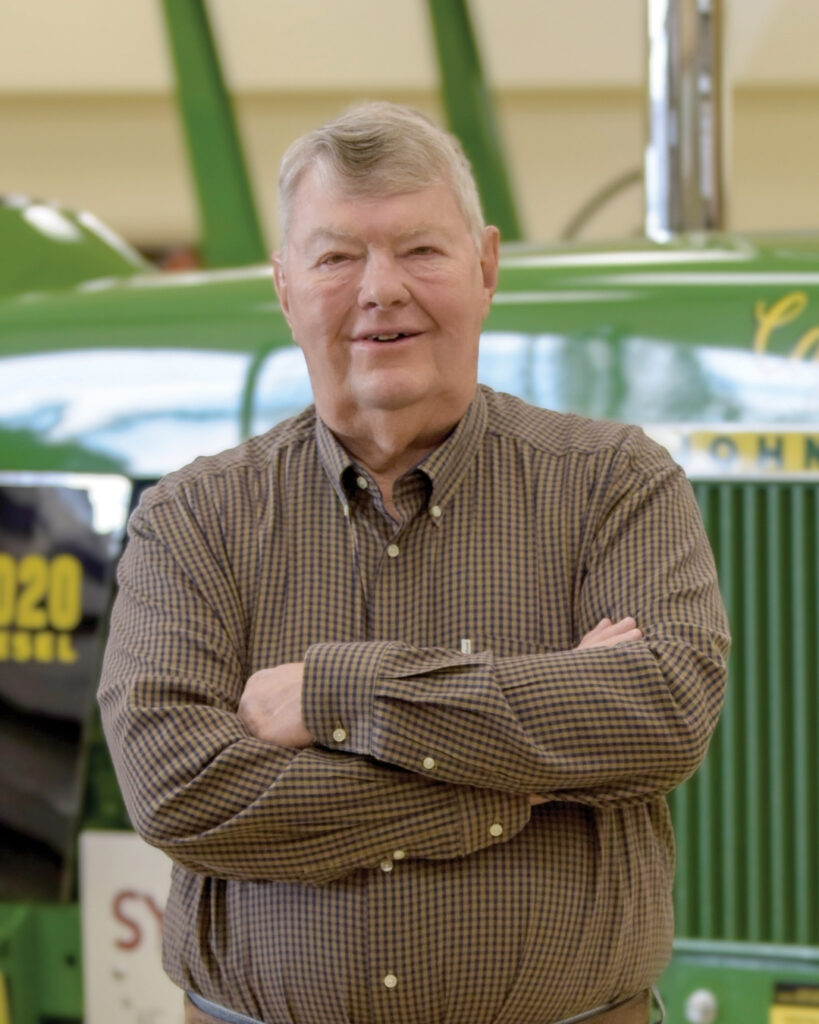 Eddie Sydenstricker with gray hair, wearing a brown plaid shirt, stands smiling with arms crossed in front of a green John Deere tractor indoors.