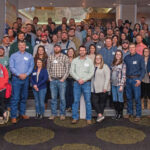A large group of people, wearing casual and semi-formal attire, are gathered indoors for a group photo. Some are standing on steps, and a few are leaning against the walls. The setting appears to be a conference or social event.