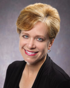 Portrait of Tami Ensor Executive assistant and Corporate board of directors secretary with short, light brown hair smiles at the camera. She is wearing a black top and small hoop earrings against a gray, textured background.