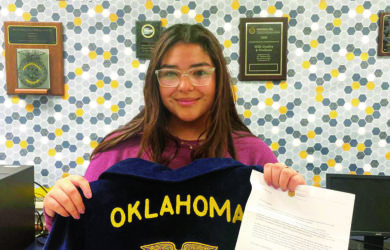 A young person wearing glasses holds up a blue jacket with Oklahoma and an emblem on it. They are standing in front of a honeycomb-patterned wall with plaques. Theyre also holding a letter in the other hand.