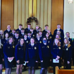 A group of people, mostly students in matching dark blue jackets, pose in rows inside a church with two large circular stained glass designs on the walls. A man in a light shirt stands to the left. The setting is bright and formal.