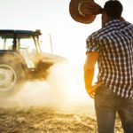 A person in a plaid shirt and jeans stands with their back to the camera, holding a straw hat. They face a tractor driving through a dusty field under a clear sky, evoking a rural farm scene.