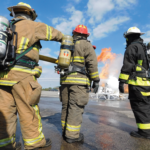 Three firefighters in gear, including helmets and oxygen tanks, stand on a wet tarmac. They face a large flame and smoke in the background against a partly cloudy sky, preparing to extinguish the fire.