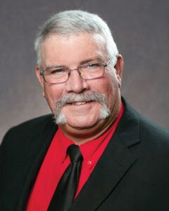 Portrait of Monte Fisher District 2 Northeast board member smiling man with gray hair and a mustache, wearing glasses. He is dressed in a black suit with a red shirt and a black tie, posed against a plain gray background.
