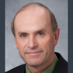 Portrait of a middle-aged person with short, thinning hair, wearing a dark suit jacket and a green shirt. The background is a neutral gray.