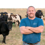 A man with crossed arms stands smiling in front of a herd of cows in a grassy field. He wears a blue t-shirt and jeans. The sky is overcast, and the cows are various colors, including black, brown, and white.
