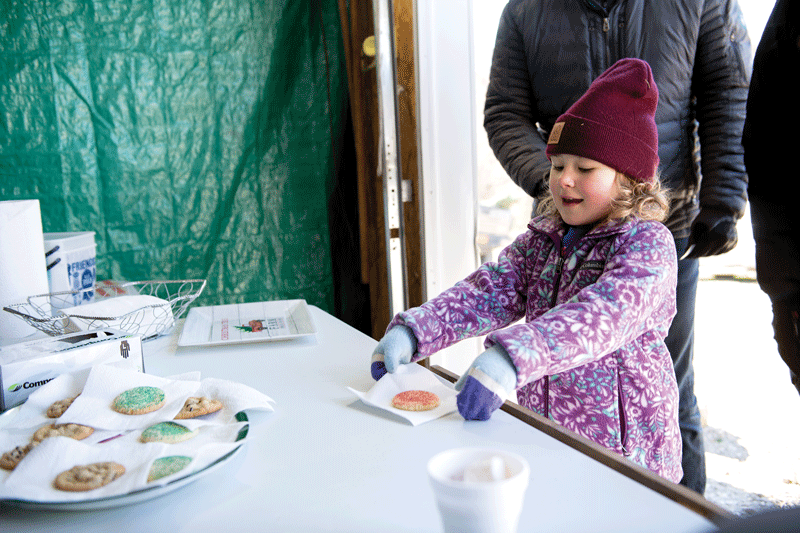A smiling child in a purple coat and maroon hat takes a cookie from a table with assorted cookies. There are two other people partially visible, and a green tarp hangs in the background.