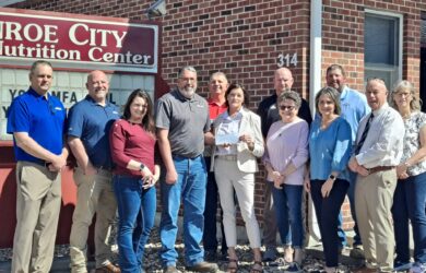 Monroe City Senior Nutrition Center stands in front of a brick building and a sign reading “Monroe City Nutrition Center.” Most are smiling and dressed in business casual attire.
