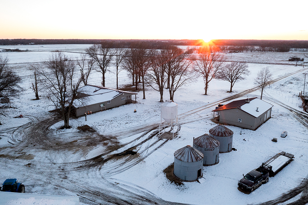 Aerial view of a snowy landscape of Gleonda Angus Farms at sunrise. Three silos, two buildings, and a truck are in the foreground. Bare trees and snow-covered fields stretch into the distance. A tractor is visible in the bottom left. The sun is setting on the horizon.