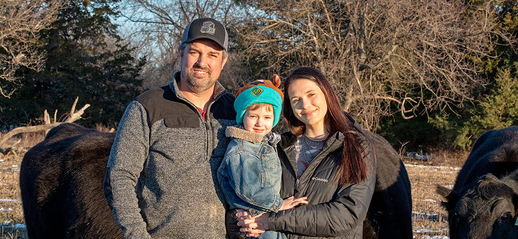 Traves and Kenadee Merrick and their son Denver stand together outdoors with cattle in the background. The Merricks are smiling, while the child wears a colorful hat and is held by the Kenadee. Bare trees and a clear sky are visible.