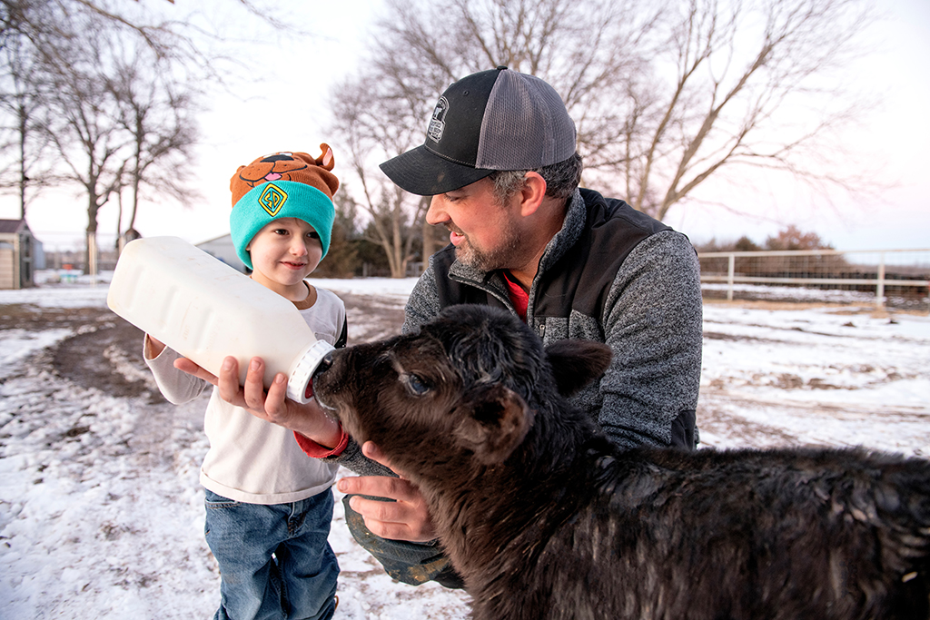 Traves Merrick crouches next to his son Denver, wearing a blue jacket and hat, holding a large milk bottle to feed a black calf. They are outdoors in a snowy area with bare trees in the background.