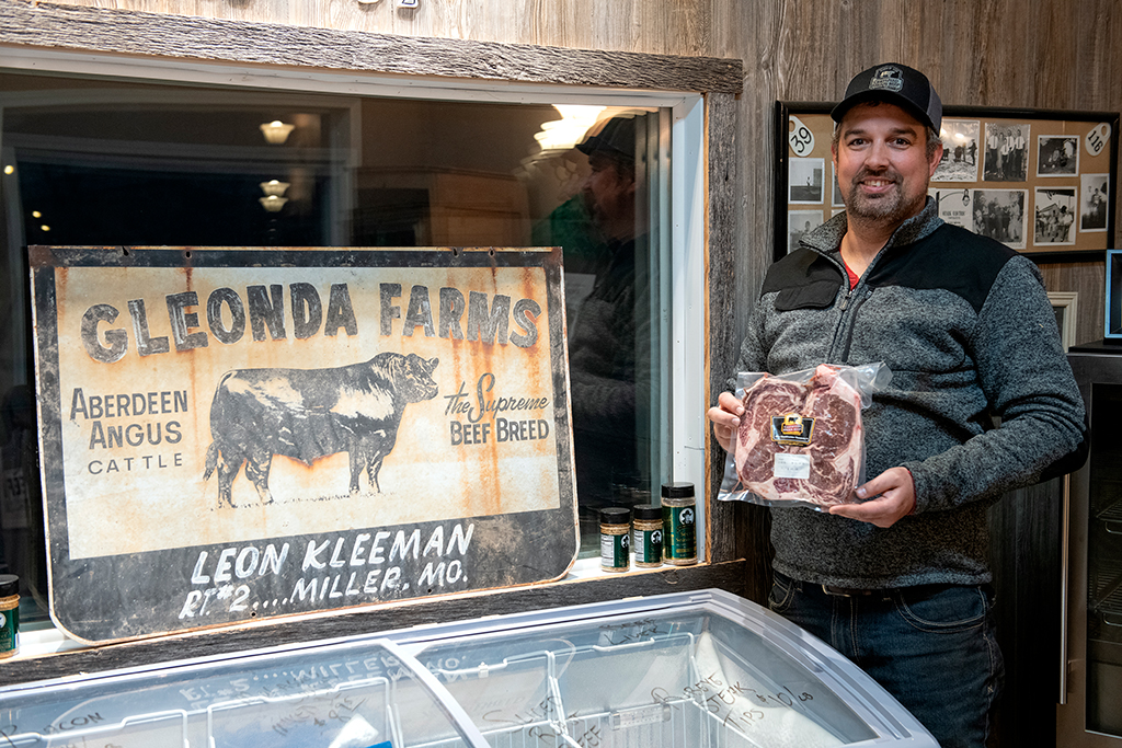 2. Traves Merrick stands holding a package of meat in front of a display case. Behind him is a vintage sign reading Gleonda Farms, Aberdeen Angus Cattle with a cow illustration. There are framed photos on the wall and spice jars near the display.