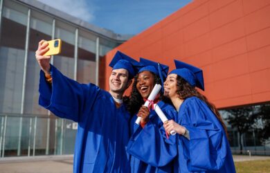 Three graduates in blue caps and gowns smile and pose for a selfie outside a modern building, each holding diplomas and celebrating their achievement.