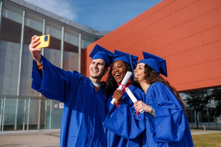 Three graduates in blue caps and gowns smile and pose for a selfie outside a modern building, each holding diplomas and celebrating their achievement.