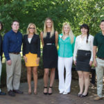 Seven people are standing in a row outdoors in a park-like setting with trees in the background. They are dressed in business casual attire, smiling at the camera.